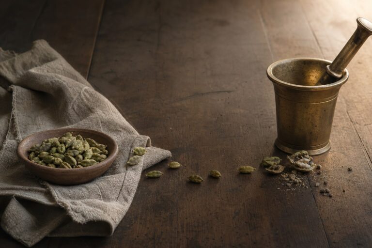 Green cardamom pods travelling across a wooden table towards a worn brass mortar