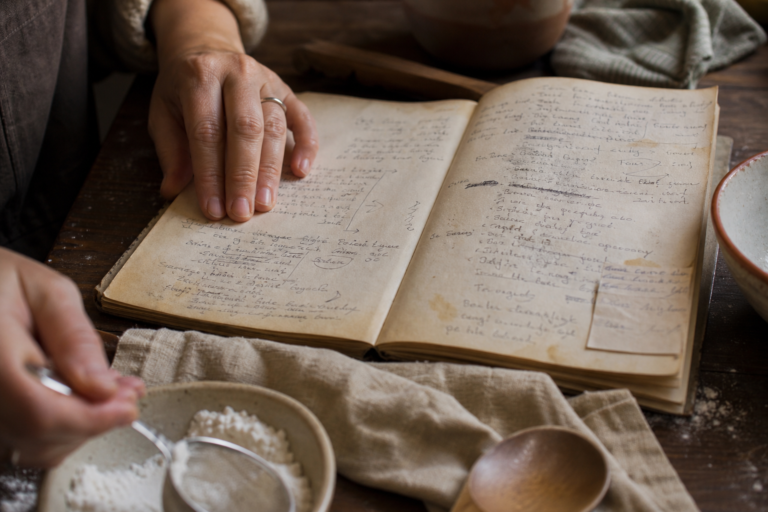 Hands consult a well-used recipe notebook while measuring flour